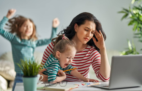 Une jeune mère travaillant à domicile sur son ordinateur portable se touche la tête de douleur alors que deux jeunes enfants s'agitent autour d'elle